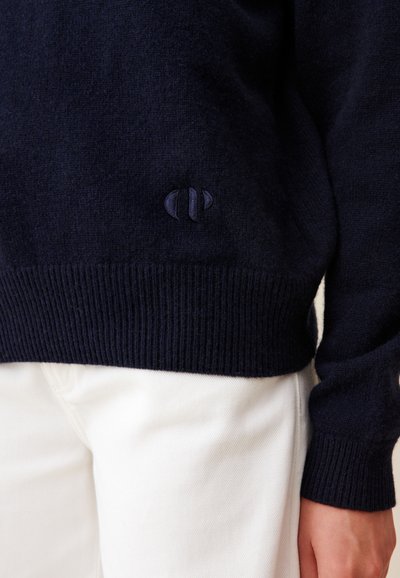 Pulover tricotat de culoare albastru închis cu tiv elastic, având un logo brodat într-o nuanță mai închisă. Asortat cu pantaloni albi.