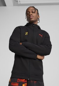 Sort zip-up hoodie lavet af blødt stof med høj krave. Har rødt Puma-logo og gult accent på venstre bryst. Glat tekstur.
