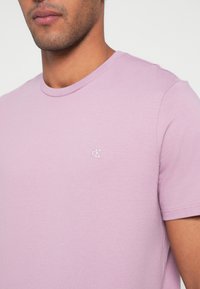 Trumpa šviesiai violetinė medvilninė marškinėliai su apvalia iškirpte ir mažu baltu logotipu ant krūtinės. Lygus paviršius ir prigludusi forma.