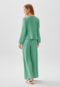 Completo verde in due pezzi con motivo a trama. Il top ha maniche lunghe e un design a peplo, abbinato a pantaloni a gamba larga e sandali bassi.