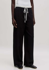 Schwarze weit geschnittene Hose aus weichem Material, mit einem wei�ßen Kordelzugbund. Getragen mit einem eng anliegenden weißen Tanktop und schwarzen Schuhen.