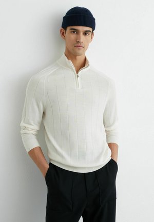 Biały prążkowany sweter na zamek z długimi rękawami i wysokim kołnierzem, zestawiony z czarnymi spodniami. Dzianinowa faktura z delikatnym pionowym wzorem w paski.