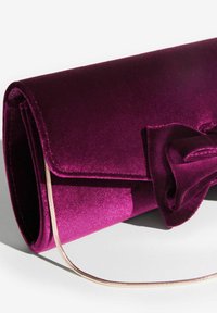 Violets samta rokassoma ar dekoratīvu loku un plānu zelta ķēdes siksnu, kas atrodas uz balta virsmas.