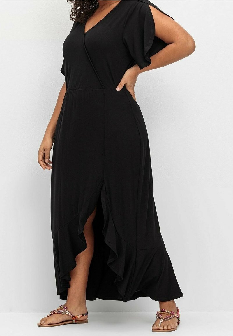 Femme en robe midi noire à col en V avec ourlet volanté et fente avant, portant des sandales plates à motifs, posant avec une main sur la hanche.