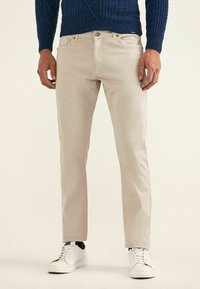 Chino en coton beige avec une coupe slim, dotés de poches avant discrètes et d'une ceinture épurée. Associés à des baskets blanches et à un pull en tricot bleu.