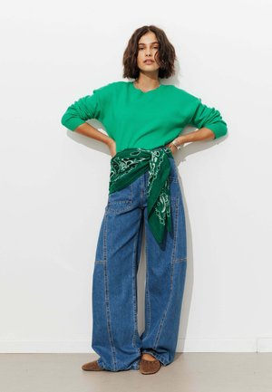 Femme aux cheveux courts et foncés portant un pull vert, un jean large bleu, des chaussures marron et une écharpe verte à motifs nouée autour de sa taille.