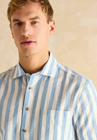 Hele sinine ja valge riba lõngast särk lühikeste varrukatega, millel on nööbid alla ja frontaalne tasku ning nööbist accents.