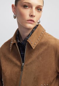 Veste en suède marron avec des clous argentés le long du col. Présente une fermeture éclair et est superposée sur une chemise à carreaux aux motifs bleus et verts.