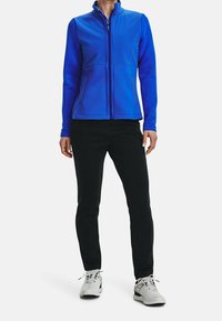 Kvinna klädd i en blå zip-up jacka, svarta byxor och vita sneakers, stående mot en helt vit bakgrund.