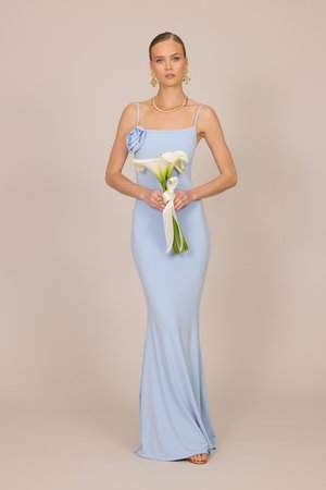 Frau in einem hellblauen, ärmellosen Kleid, die weiße Calla-Lilien hält, goldene Ohrringe und eine Halskette trägt, steht vor einem einfarbigen Hintergrund.