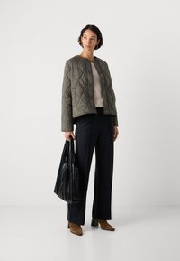 Vrouw in een khaki gewatteerde jas, beige sweater, zwarte wijde broek, bruine heteluchtschoenen, met een grote zwart gestreepte tote bag.
