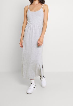 Kobieta ubrana w jasnoszara bezrękawnikową sukienkę maxi z rozcięciem po boku oraz białe sneakersy z czarnym logo Swoosh.
