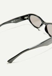 Black sunglasses with a smooth finish, oval lenses, and a sleek design. Features a hinge on the temple and includes CE and UKCA markings.