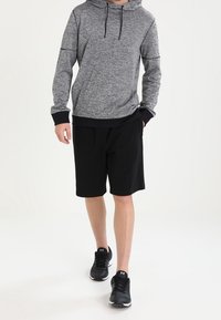 Osoba ubrana w szary hoodie, czarne szorty i czarne buty sportowe idzie na białym tle.