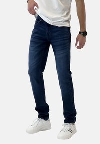 Jeans de mezclilla oscura con corte ajustado, que presentan un sutil patrón de desgaste. Combinados con zapatillas blancas y una camiseta blanca lisa.