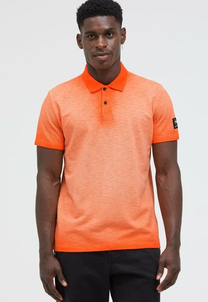 Mann, der ein orangefarbenes kurzärmeliges Polohemd mit schwarzen Knöpfen und schwarze Hose trägt, steht vor einem schlichten Hintergrund.