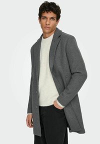 Grau Wollmantel mit einer maßgeschneiderten Passform, schmalen Revers und zwei Knöpfen. Über einem cremefarbenen Strickpullover getragen und mit schwarzen Hosen kombiniert.