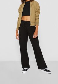 Khaki bomberka na czarnym topie cropped, zestawiona z luźnymi czarnymi spodniami cargo i czarnymi sneakersami z białą podeszwą. Casualowy strój.