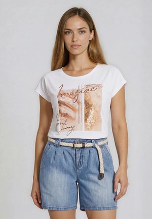 Kobieta z długimi prostymi włosami, ubrana w biały t-shirt z brązowym napisem i dżinsowe szorty z plecionym beżowym paskiem, stojąca na szarym tle.