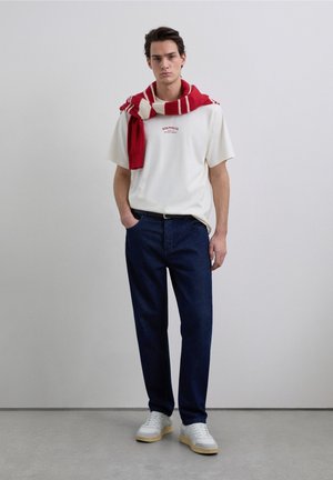 Wit T-shirt met rode tekst, donkerblauwe spijkerbroek, grijze sneakers met beige zolen, en een rood-wit gestreepte sjaal om de nek gedrapeerd.