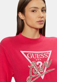 Sudadera rosa con escote redondo, que presenta un logo triangular blanco y embellishments de rhinestone que dicen "Amor". Textura de tela suave.
