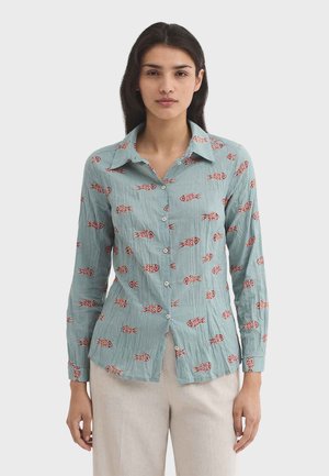 Woman wearing a light blue long-sleeve button-up shirt with red fish patterns and beige pants, standing against a plain background.