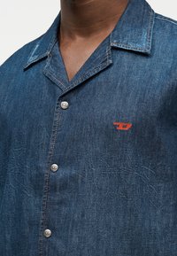 Κοντινό πλάνο ενός ατόμου που φοράει σκούρο μπλε πουκάμισο από τζιν με γιακά κάμπινγκ, ασημένια κουμπιά και μικρό κόκκινο κεντημένο λογότυπο στο στήθος.