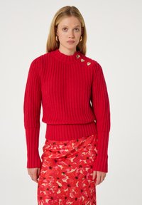 Roter Rippstrickpullover mit hohem Kragen, der mit drei goldenen Herz-Akzenten an der Schulter versehen ist, kombiniert mit einem floralen rosa Rock.