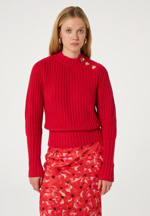 Rode geribbelde trui met een hoge halslijn, versierd met drie gouden hartaccenten op de schouder, gecombineerd met een bloemenrok in het roze.