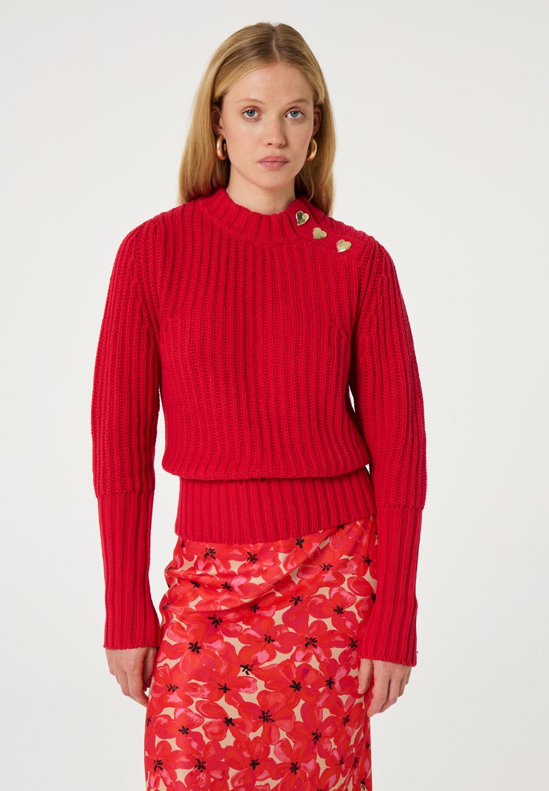 Roter Rippstrickpullover mit hohem Kragen, der mit drei goldenen Herz-Akzenten an der Schulter versehen ist, kombiniert mit einem floralen rosa Rock.