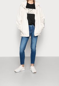 Puffig cremefärgad jacka med huva, svart grafisk sweatshirt, blå skinnyjeans och vita sneakers. Släta texturer och minimala detaljers användning av hårdvara.