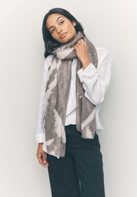 Sciarpa grigia e crema con motivi astratti e sottili accenti metallici, realizzata in un tessuto morbido e strutturato, elegantemente drappeggiata intorno al collo.