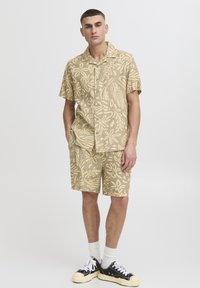 Lühikeste varrukatega beež särk ja sobivad lühikesed püksid valge lehemustriga, valmistatud kergest materjalist. Mustad tossud valgete paeltega.