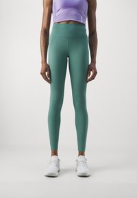 Ett par högmidjade, figurnära leggings i en enfärgad grön nyans, tillverkade av ett mjukt och stretchigt material, i kombination med en ljuslila sportbehå.