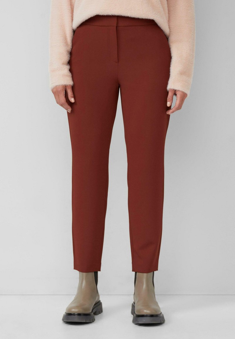 Persoon in roestkleurige slim-fit broek, beige chunkybots, en een lichtbeige fuzzy trui die tegen een effen achtergrond staat.