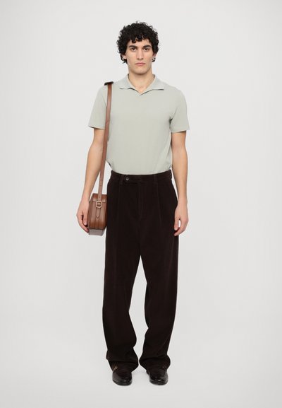 Heledhall hall polo särk, tumepruunid sametpüksid, pruun naha postbag, mustad libisevad kingad, seismine valge tausta vastu.
