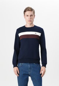 Marineblå sweatshirt med rund hals, prydet med horisontale striper i hvit og burgunder. Inkluderer grønn logo detalj på brystet.