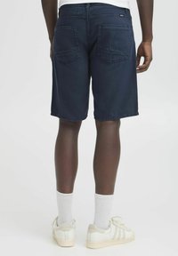 Navy blue denim shorts with a straight cut, featuring two back pockets and stitched detailing. Worn with white sneakers and socks.