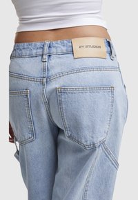 Personne portant un jean bleu clair avec des poches arrière visibles et une étiquette beige 2Y Studios, associé à un haut court blanc.