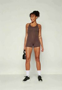 Woman wearing brown sleeveless workout romper holding a black kettlebell, standing and smiling against a plain light background.