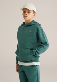 Vihreä huppari, jossa on kengurupocket ja musta logo rinnassa; yhdistetty vastaaviin housuihin, ja käytetty valkoisen pesäpallolakin kanssa.