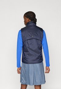 Colete azul-marinho sem mangas feito de material leve, com gola alta e textura suave. Por baixo, vê-se uma blusa azul de mangas compridas acompanhada de uma saia azul com padrão.