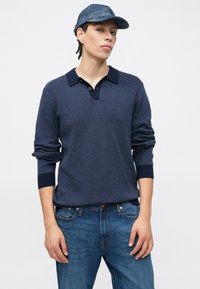 Marineblauer Strickpullover mit Kragen und zwei Knöpfen, kombiniert mit blauen Jeans. Das Modell trägt eine blaue, gemusterte Mütze. Lässiger Look mit gerippten Bündchen.