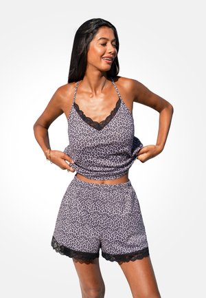 Vrouw die een paarse camisole met luipaardprint en bijpassende shorts met zwarte kanten rand aanpast, staand tegen een egale lichte achtergrond.