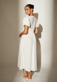 Weißes Midikleid mit Puffärmeln und taillierter Taille. Es verfügt über einen fließenden Rock und eine glatte Textur. Kombiniert mit beigen Sandalen.