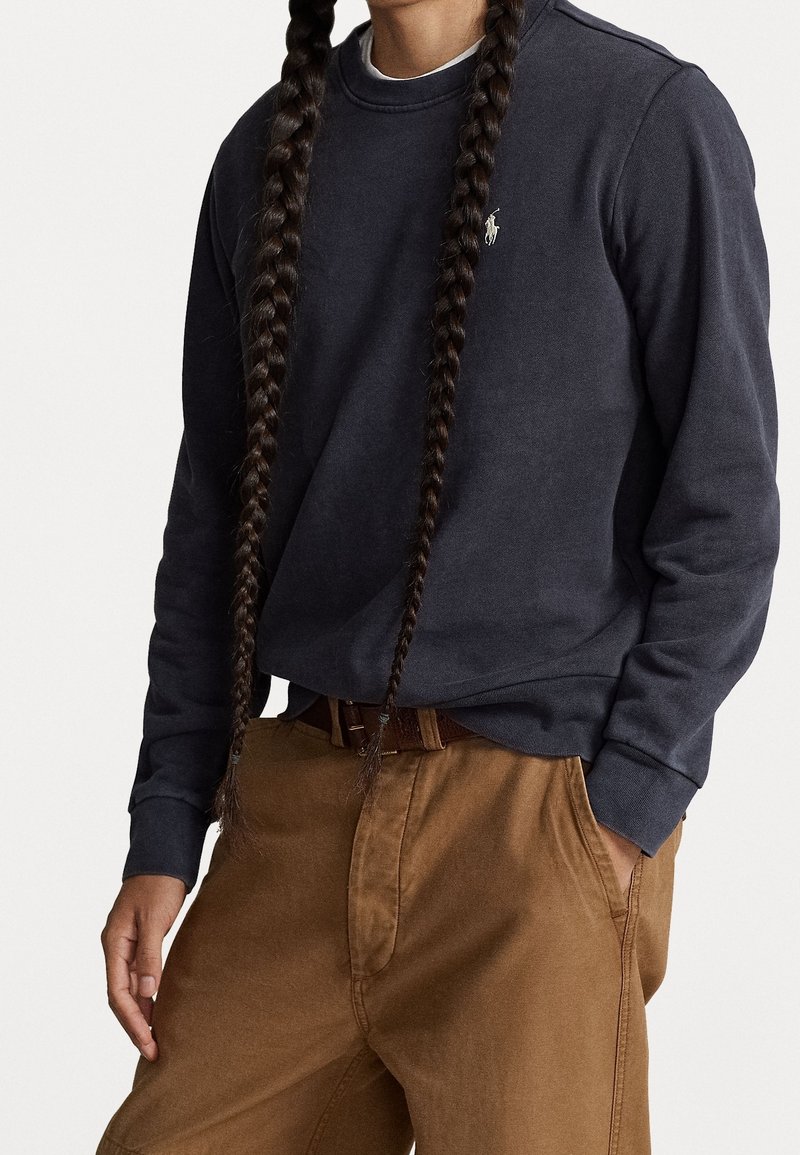 Dunkelblaues Sweatshirt mit Rippbündchen und Rundhalsausschnitt, mit kleinem gesticktem Logo; kombiniert mit braunen, locker sitzenden Hosen.
