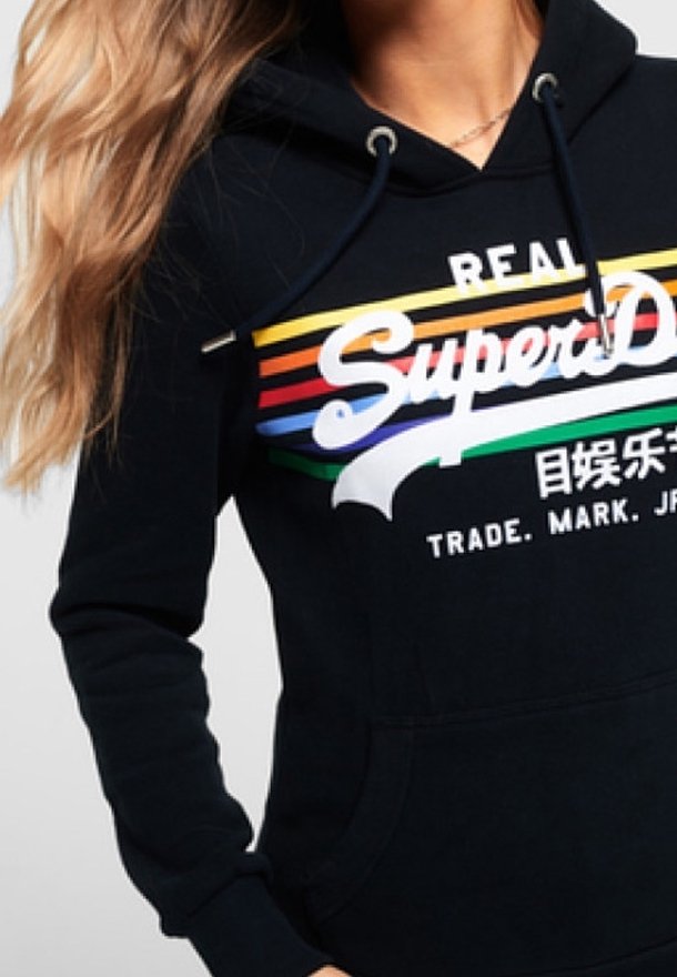Donna che indossa un hoodie nero con strisce orizzontali colorate e il testo bianco "SuperDry" sul petto.