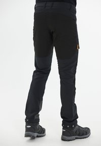 Pantalons de plein air noirs avec un design en matériaux mixtes, accents en maille, poches zippées et coupe fuselée. Portés avec des chaussures de randonnée noires.