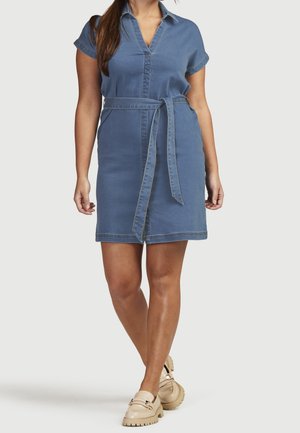 Mujer con un vestido corto de denim azul de manga corta con cinturón y mocasines beige gruesos, de pie frente a un fondo liso.