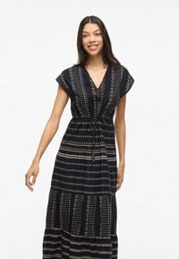 A woman with long dark hair smiles, wearing a black patterned dress with short sleeves and a cinched waist, against a light background.